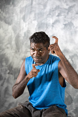 A man in a blue sleeveless top making hand gestures against a textured grey background.