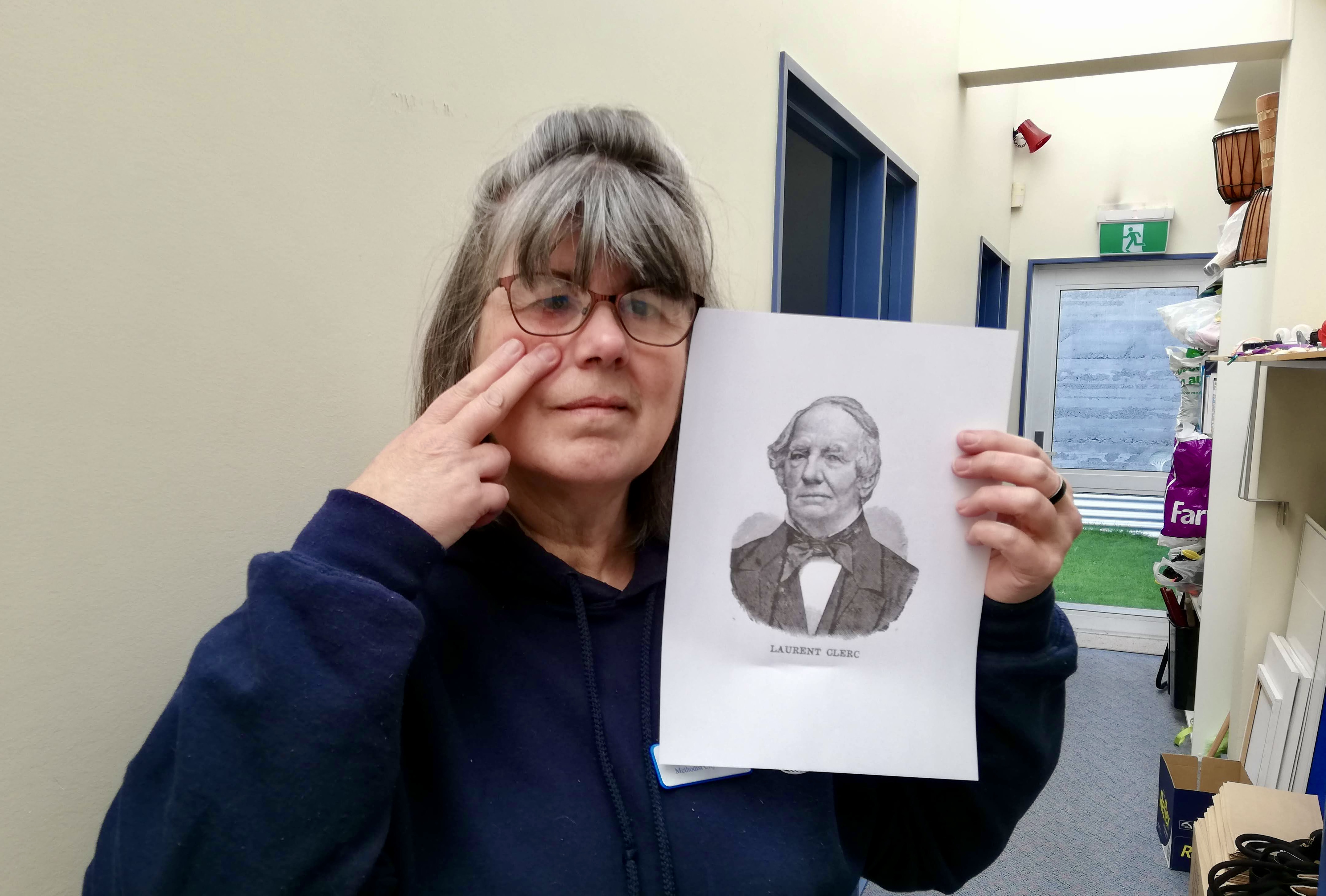 A woman with glasses holds a printed portrait of Laurent Clerc while pointing to her cheek in a hallway.