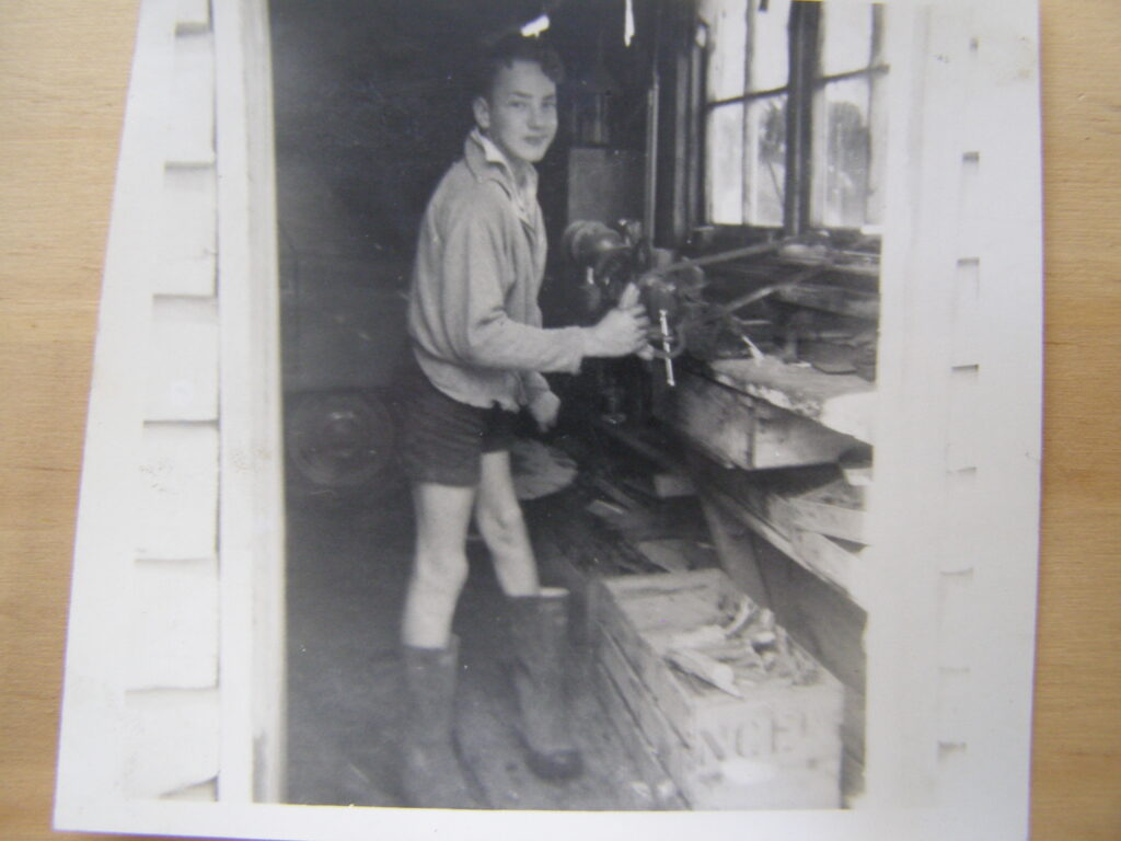 A young boy standing in a workshop, holding a tool and wearing shorts and boots, with wooden shelves and windows in the background.