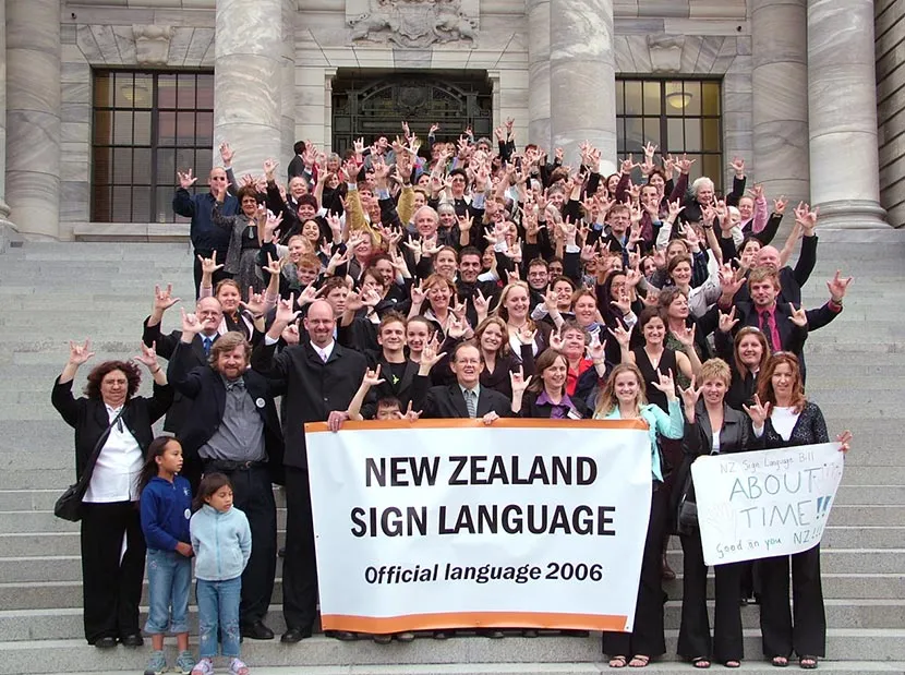 A large group of people celebrating New Zealand Sign Language, holding up their hands in the sign for 'I love you' in front of a historic building, with a banner declaring New Zealand Sign Language as an official language.