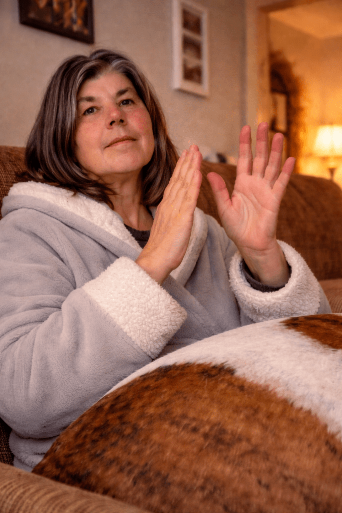 A woman in a cozy grey bathrobe sits on a couch, with her hands raised as if demonstrating or explaining something, while a patterned blanket covers her lap.
