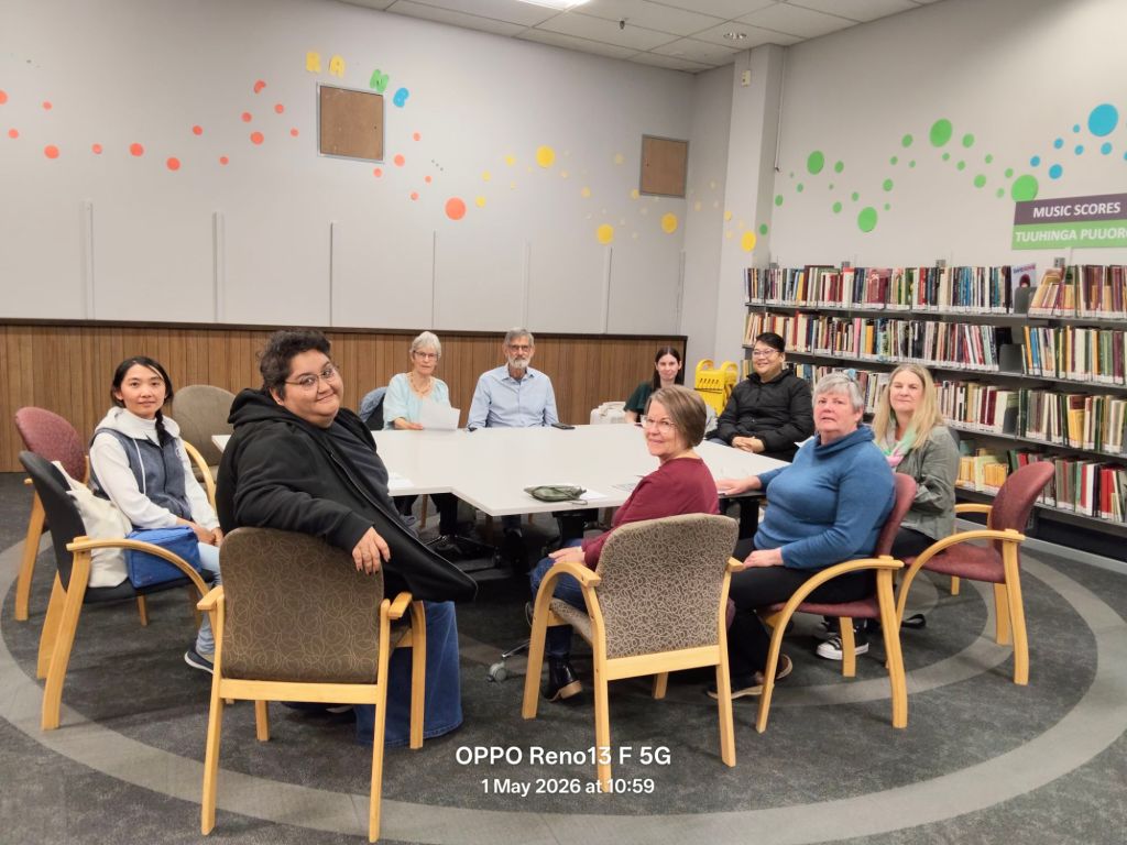 A group of diverse individuals seated around a large table in a library setting, with bookshelves in the background and colorful decorations on the walls.