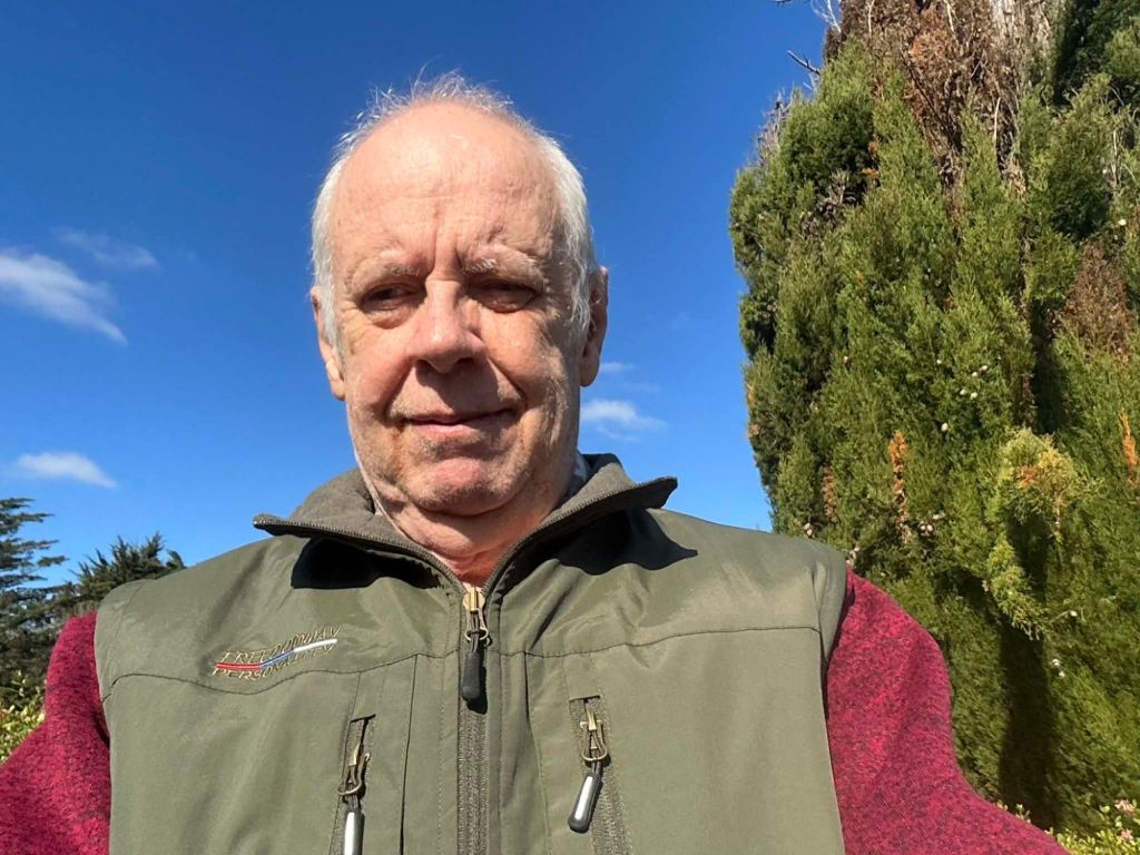 A man wearing a green vest and red sweater stands outdoors, smiling at the camera, with trees and a clear blue sky in the background.